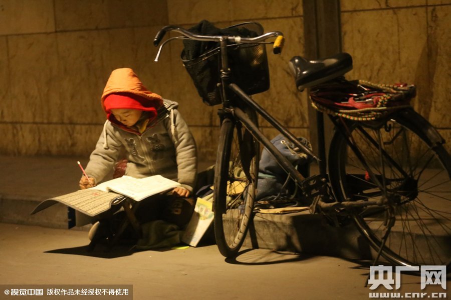 鄭州中原路京廣鐵路大橋下涵洞的馬路牙子邊，一位戴著紅色辣椒帽的小男孩趴在一個小馬扎上，上面鋪著一張車墊，在微弱的燈光下寫作業(yè)